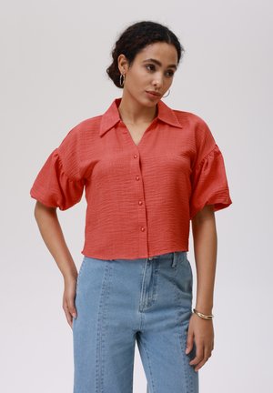 Short-sleeve, coral blouse with a button-up front and large collar. Textured fabric and relaxed fit, paired with light blue high-waisted jeans.