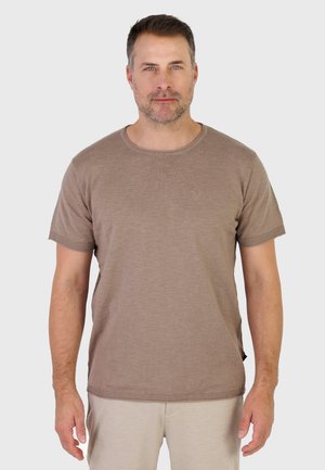 Hombre con cabello corto y oscuro y barba incipiente, lleva una camiseta de punto marrón de manga corta y pantalones beige claro, de pie contra un fondo blanco.