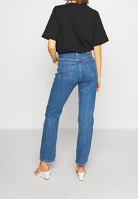 Vaqueros de mezclilla azules con corte recto y un sutil desgaste, combinados con una camiseta negra de gran tamaño. Cuenta con bolsillos traseros y un parche de cuero con el logo.
