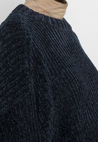 Granatowy sweter o fakturze, z prążkowanym wzorem, wysokim kołnierzem i miękką tkaniną. Sylwetka swetra jest luźna, z opuszczonymi ramionami.