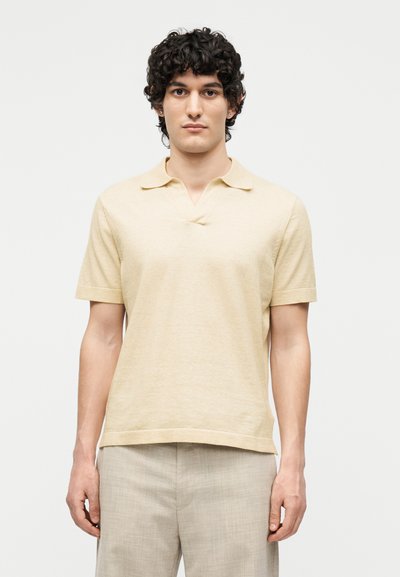 Jeune homme aux cheveux bouclés et foncés, portant un polo beige à manches courtes et un pantalon gris clair, debout devant un fond blanc uni.