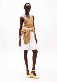 Tan mouwloos topje, witte knie-lange rok met split, en gele sleehakken. Een gebreid beige vest is om de taille geknoopt.