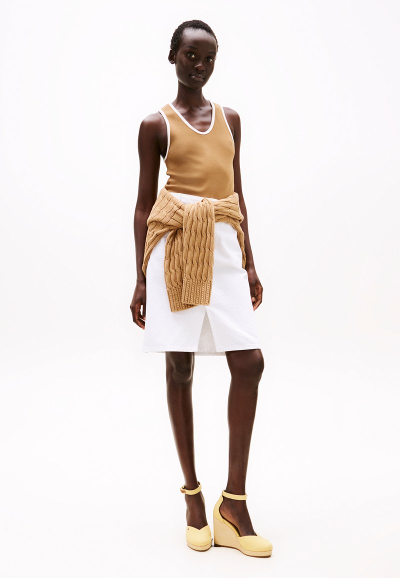 Tan mouwloos topje, witte knie-lange rok met split, en gele sleehakken. Een gebreid beige vest is om de taille geknoopt.