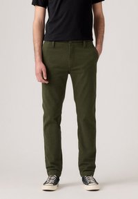 Man wearing olive green trousers, black t-shirt, and black-and-white sneakers, standing with one hand in pocket against plain background.