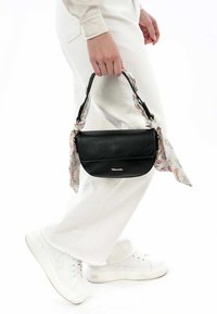 Black leather handbag with a curved flap, featuring a floral-patterned scarf tied around the handle. Model wearing white jeans and sneakers.