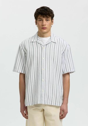 Jeune homme portant une chemise blanche à manches courtes avec des rayures verticales grises et un pantalon beige, debout devant un fond uni.