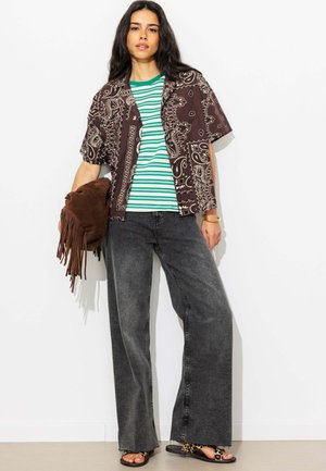 Femme portant une chemise marron à manches courtes à motifs sur un top rayé vert, un jean large et foncé, des sandales imprimé léopard et tenant un sac marron à franges.