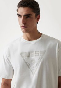 Hvid bomulds T-shirt med et stort, afdæmpet trekantet logo med "GUESS" og tekst. Rund hals og korte ærmer. Glat tekstur.