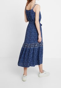 Robe maxi bleue à imprimé floral avec un corsage froncé, de fines bretelles, et une jupe à étages avec un bord en dentelle. Associée à des baskets blanches.