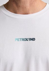 Tricou alb din bumbac cu logo-ul "PETROLIND" brodat în albastru și negru pe piept, cu un gât standard rotund și mâneci scurte.