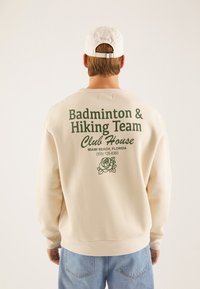 Kremfarget hettegenser med grønn tekst som sier "Badminton & Hiking Team" og en rosegrafikk. Sammen med lyseblå jeans og en hvit caps.