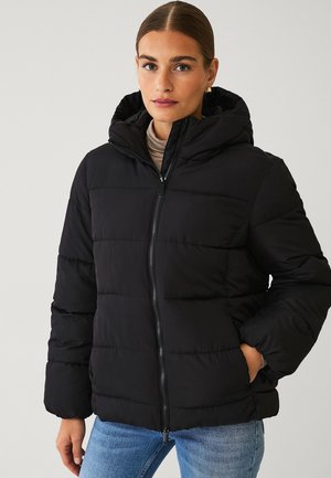 Frau trägt eine schwarze Steppjacke mit Kapuze, einen beigen Rollkragenpullover und blaue Jeans und steht vor einem schlichten hellen Hintergrund.