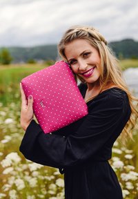 Žena s dlouhými blond vlasy v černých šatech se usmívá, když drží růžovou psaníčko s puntíky venku s zelení a květinami v pozadí.