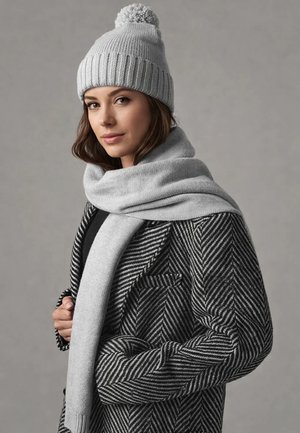 Frau trägt eine graue Strickmütze mit Bommel, einen passenden Schal und einen schwarz-grauen Fischgrätenmantel, posiert vor einem einfarbigen grauen Hintergrund.