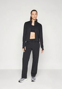 Naine seisab, seljas must tõmblukujope, must spordirinnahoidja, mustad püksid ja hallid sportlikud jalanõud, lihtsa heleda tausta taustal.