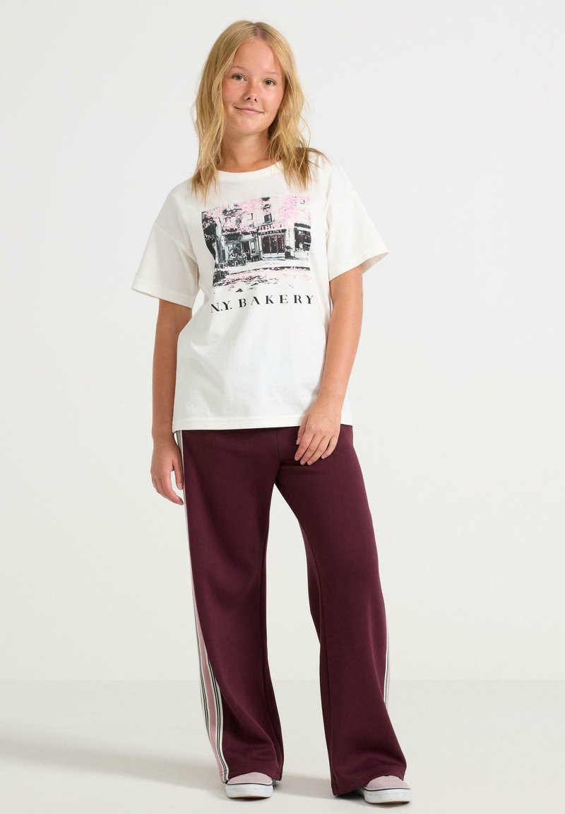 Baumwoll-T-Shirt in Off-White mit einem schwarzen Grafikaufdruck mit der Aufschrift "N.Y. BAKERY", kombiniert mit kastanienbraunen Sweatpants mit weißen Seitenstreifen.