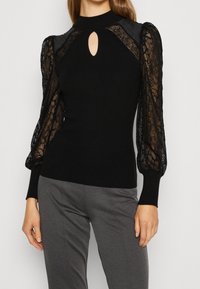 Haut en tricot noir avec des manches volumineuses en dentelle, un col montant et un petit détail en forme de trou de clé au niveau de la poitrine. Le tissu présente une texture côtelée.