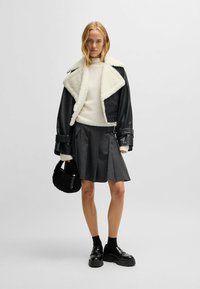 Schwarze Lederjacke mit weißem Lammfellkragen, cremefarbener Pullover, grauer plissierter Rock, schwarze klobige Schuhe und schwarze runde Tasche.
