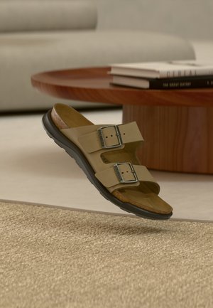 Two-strap tan sandal with metal buckles hovering above a woven beige rug, with a wooden coffee table and books in soft focus background.