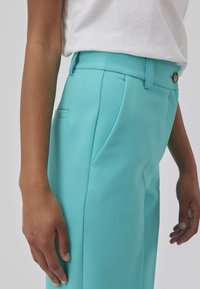 Pantalons taillés turquoise avec poches latérales, une taille plate et une texture lisse. Comprend une fermeture à bouton et des passants de ceinture à la taille.
