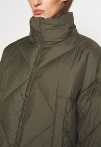 Nafukovací olivově zelená bunda s vysokým límcem, diagonálním prošíváním a předním zipem. Má boční kapsu a hladký lesklý povrch.