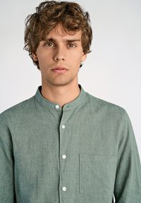 Camisa verde de botones con cuello tipo banda, fabricada en lino texturizado, con un bolsillo delantero y botones blancos, llevada por un modelo con pelo rizado.