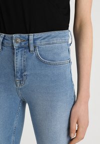 Jean en denim bleu clair à taille haute, avec une poche avant, fermeture à bouton en métal et texture lisse. Les coutures sont visibles.