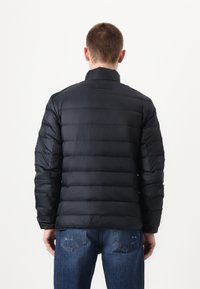 Giubbotto imbottito nero con design a righe orizzontali, colletto alto e una texture liscia e lucida. Indossato con jeans blu.