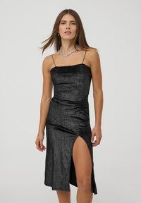 Abito nero glitterato con silhouette aderente, spalline sottili e uno spacco laterale. Il tessuto ha una finitura lucida e testurizzata.