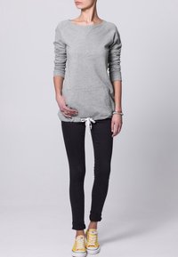 Sudadera gris con mangas largas, cuello redondo y bolsillos frontales, combinada con jeans ajustados negros y zapatillas amarillas.