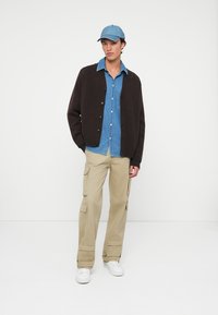 Brun kofta, blå denimskjorta, beige cargobyxor med fickor, vita sneakers och en blå keps. Enkelt mönster, avslappnad passform och lager-på-lager-stil.