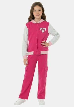 Fille debout, main sur la hanche, portant une veste à capuche rose et grise et un pantalon cargo rose assorti avec des baskets blanches.