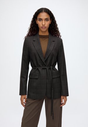 Femme aux cheveux bouclés porte un blazer ceinturé à carreaux foncés sur un haut marron et un pantalon marron ample sur un fond clair uni.