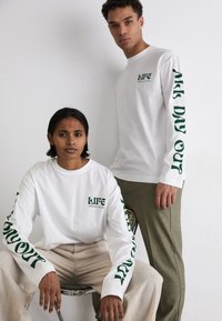 Weiße Langarm-T-Shirts mit grünem Drucktext, auf dem die Wörter "LIFE" und "ALL DAY OUT" auf den Ärmeln stehen. Ein Modell in beigen Hosen sitzt auf einem Stuhl.