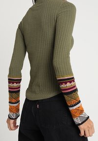 Slim-fit olivgrön ribbad tröja med mångfärgat mönstrade ärmar, bärs av person med svarta denimjeans, visad bakifrån.