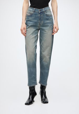 Person iført falmede blå jeans med høj talje og sorte ankelstøvler med spids tå, stående mod en ensfarvet lys baggrund.