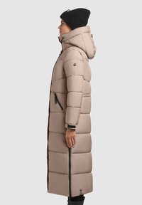 Világos bézs hosszú steppelt kabát kapucnival, párnázott részekkel, oldalzippel és fekete részletekkel, fekete sapkával viselve.