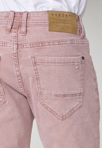 Roze traperice s visokim strukom, s kožnom oznakom branda, dvostrukim stražnjim džepovima i suptilnim šavnim detaljima.