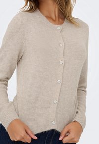 Cardigan beige en maille avec encolure ronde, ourlet et poignets côtelés, fermé par six boutons décoratifs en forme de fleur.