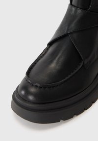 Botină din piele neagră, cu vârful rotund, având un design cu curea încrucișată, cusături vizibile și o talpă din cauciuc texturat.