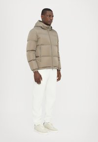 Puffer jacket in muted beige, featuring a hood and zip front, with horizontal stitching. Model wears white pants and light sneakers.