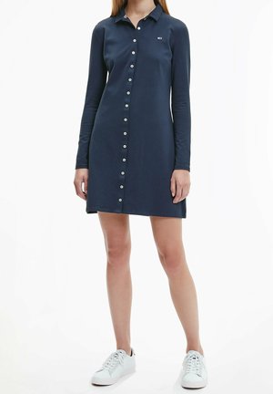 Woman wearing a navy blue long-sleeve button-down dress and white sneakers, standing against a plain white background.