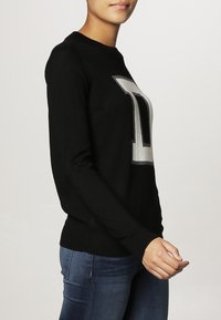 Black sweater featuring a large silver letter "D" on the front, crew neck, and long sleeves. Smooth texture, slim fit.