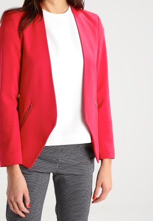 Femme portant un blazer rouge vif sur un haut blanc et un pantalon à carreaux noir et blanc, avec les ongles peints en rouge.
