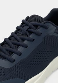 Αθλητικό παπούτσι σε ναυτικό μπλε χρώμα με υφασμάτινη πλέξη, χονδρά κορδόνια, λείες επενδύσεις και λευκή σόλα, εμφανιζόμενο σε κοντινή γωνιακή λήψη.