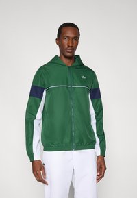 Zielona kurtka z kapturem z granatowymi i białymi ukośnymi akcentami, lekki materiał, zapięcie na zamek, elastyczne mankiety i małe logo na piersi.