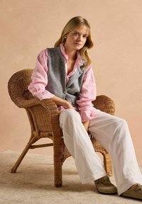 Grau gestrickte Weste über einem rosa-weiß gestreiften Hemd, kombiniert mit weißen Leinenhosen. Sitzt in einem geflochtenen Stuhl auf einer strukturierten Oberfläche.