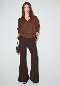 Suéter marrón de cuello en V con corte holgado, combinado con pantalones de campana oscuros. Accesorios incluyen un pequeño bolso texturizado y una pulsera ancha.