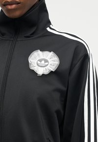 Person trägt eine schwarze Jacke mit Reißverschluss, weißen Streifen an den Ärmeln und einem weißen, floralen Mesh-Accessoire mit einem Logo auf der Brust.