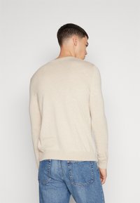 Pulôver bege de manga longa com acabamento texturizado, punhos e bainha canelados. Usado com jeans de denim azul, apresentando um ajuste solto e um estilo casual.